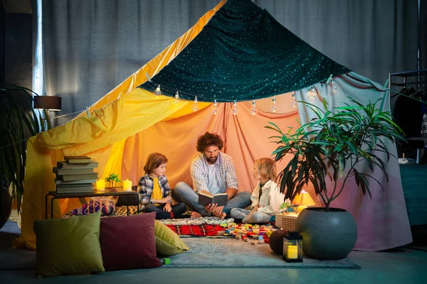 A very happy dad is having a good time with his two little boys as they are all reading a book in a blanket indoor tent surrounded by many pillows. Family