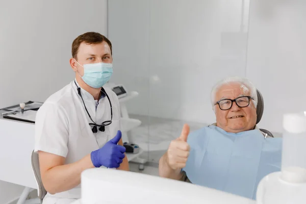 歯医者のオフィスのカメラの前で医者と患者はカメラの前でポーズカリスマ老人患者は歯医者の椅子の上に寝そべって医者は保護マスクを着て — ストック写真