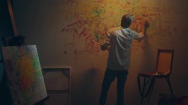 Büyük sanat stüdyosunda ressam duvara yeni bir resim çiziyor yağlı boya paletini elinde tutuyor ve parmaklarıyla resmi boyuyor. 4k