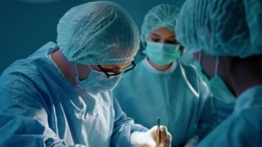 Bir erkek cerrah, ameliyat odasında iki asistanından yardım alırken hastasını ameliyat etmek için cımbız kullanıyor..