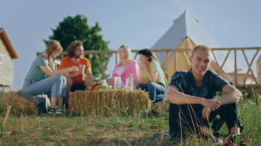 İyi görünümlü Afro-Amerikan bir adam kameraya bakıyor ve kamp alanının ortasında çimlerin üzerinde otururken poz veriyor ve arka plandaki arkadaş grubunda doğada çok ırklı bir ürperti ve