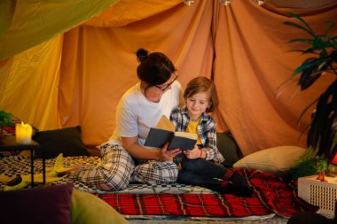 Bir annenin oğlunun yanında battaniye kalesinde oturan oğluna masal kitabı okurken daha yüksek bir kamera görüntüsü. Aile
