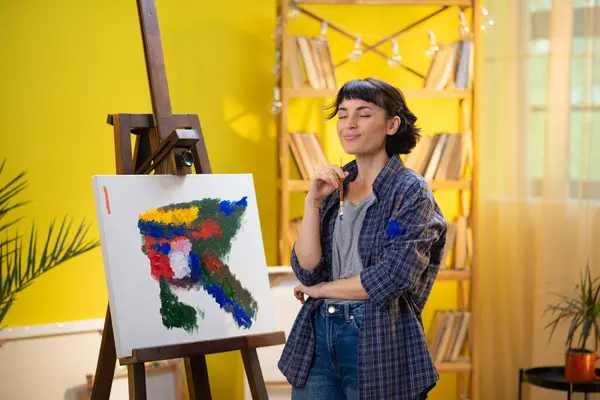 A woman is rustling through many different types of brushes while painting something astonishing on a large canvas that is held up by an easel.
