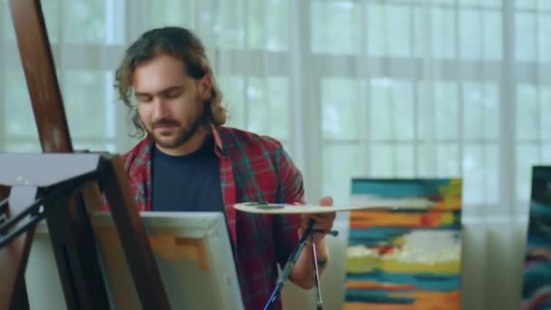 男のアーティストは 色のパレットペイントブラシを抱きながらキャンバスを描き終え 自分の作品を賞賛する彼のキャンバスに向かって探しています Arri Alexaで撮影する — ストック動画