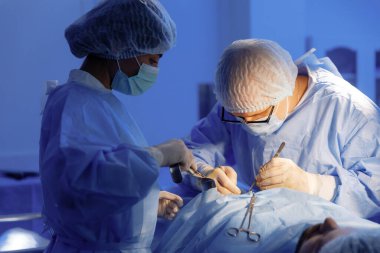 Tam teşekküllü bir cerrah üniforması giyen bir erkek doktor yoğun bir ameliyat gerçekleştirirken, her iki tarafındaki asistanları da ona yardım ediyor..