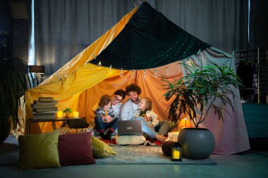 Kameranın önünde mutlu ebeveynler ve çocukları laptopta birlikte film izlerken oturma odasındaki çadırın içinde oturup patlamış mısır yiyorlar.