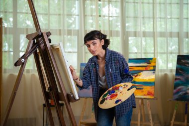 Yaratıcı genç kadın ressam projesi için yeni bir sanat çalışması üzerinde çalışıyor ve resim stüdyosunda poz veriyor. Resimlerinin önünde duran neşeli bir ressam. Boya