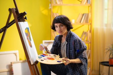 Güzel bir kadın ressam parmaklarıyla bir şey çerçevelemeye çalışıyor ve üzerinde çalıştığı tuvale boyuyor ve çok rahatlatıcı görünüyor. ressam