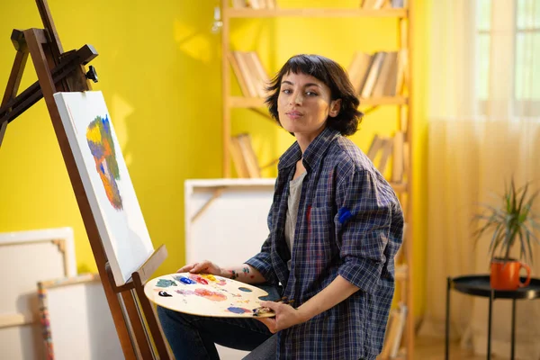 A woman seems very focused on her work as she is painting something on a canvas and wearing a blue flannel shirt with paint stains all over it. painter