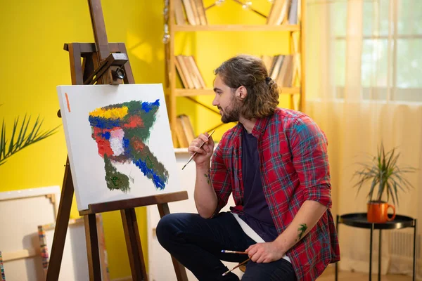 A very good looking man is painting a rare and modern painting and he is using his hands to frame something in front of him so he can paint while also being surrounded by paintings behind him. painter
