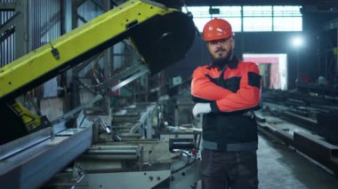 Endüstriyel imalatın ortasında dikilirken kameranın önünde poz veren koruyucu miğferi ve üniforması olan yakışıklı bir fabrika işçisi. 4k