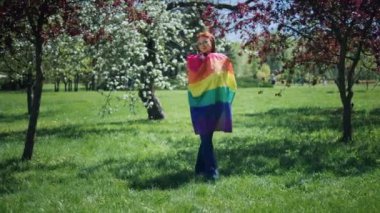 Mükemmel güneşli bir gün lgbt bayrağını tutan ve kameraya parkın ortasındaki çimlerin üzerinde duran bayan. 4k