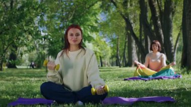 Parkın ortasında egzersiz yapan iki bayan ağırlık ve yoga minderlerini alır ve kameranın önündeki formda vücut egzersizleri için çimlerin üzerinde dururlar. 4k