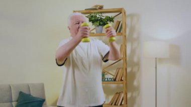 Ev antrenmanı ve formda kalma kavramı çok hevesli yaşlı bir adam gülümsüyor ve evinde ağırlıklarla egzersiz yapıyor. 4k