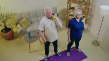 Evde birlikte yaşayıp egzersiz yapan yaşlı bir çift. Ağırlıklarıyla birlikte yoga minderinde oturan pazıları çalıştırıyorlar..