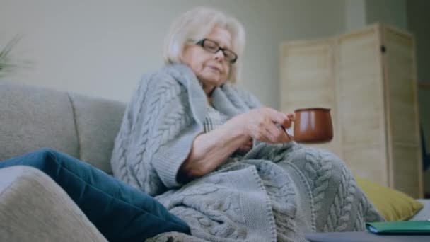 老太太坐在沙发上 拿起毛毯盖住茶具 一边喝茶 一边穿上衣服 — 图库视频影像