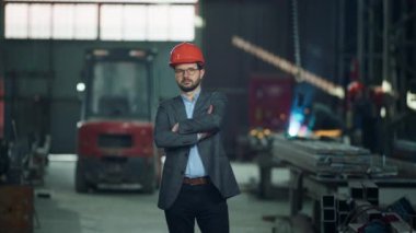 Endüstriyel fabrikadaki mühendis kameranın önünde el ele tutuşarak duruyor ve kaskını takıyor..