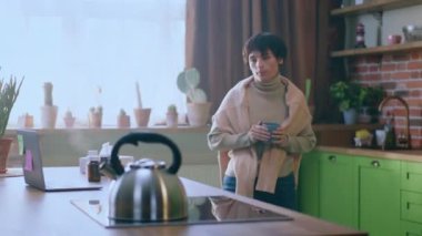 Sabahları güzel görünümlü bir kadın mutfak adasının yanında duruyor ve sıcak bir içecek yapmak için elektrikli ocaktan su ısıtıcısını almaya hazırlanıyor. 4k