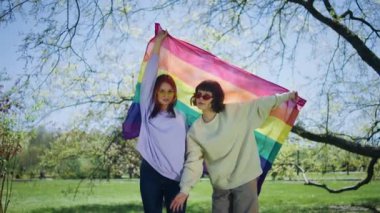 Heyecanlı lgbt çifti kameranın önünde poz veriyor ellerinde gökkuşağı lgbt bayrağı ve parkın ortasına bakıyor..