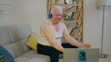 Evde oturan yakışıklı yaşlı adam kanepede oturuyor ve dizüstü bilgisayarında egzersiz yaparken bisepsleri ağırlık ile eğitiyor. 4k