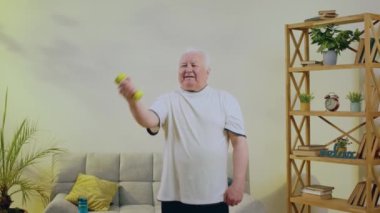 Geniş, ferah oturma odasında çok mutlu ve heyecanlı yaşlı bir adam egzersizin sonunda ağırlıkları kullanarak pazıları çalıştırmak için evde egzersiz yapıyor. 4k