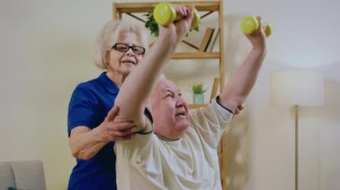 Oturma odasında oturan yakışıklı yaşlı kadın, kocası sandalyede otururken birlikte egzersiz yaparak pazıları ağırlık ile çalıştırıyor. 4k