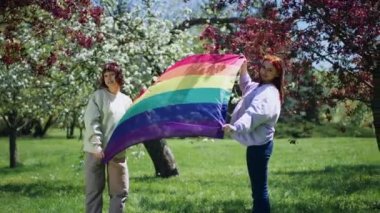 Parkın ortasında birbirine aşık olan iki genç lgbt kızı kameranın önünde Igbt bayrağını tutuyorlar. 4k