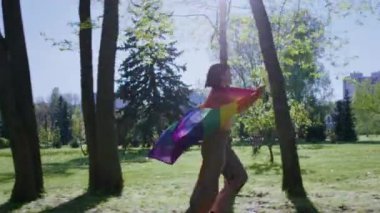 Güneşli bir günde, kısa saçlı, genç ve lezbiyen görünümlü bir kadın parkta sandalyeye otururken elinde lgbt bayrağı tutuyordu. Güneşe bakıyordu. 4k