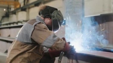 Metal fabrikasındaki profesyonel kaynakçı metal kaynak yapıyor. Güvenlik ekipmanı ve kask takıyor. Parıltılar her yere saçılıyor. 4k