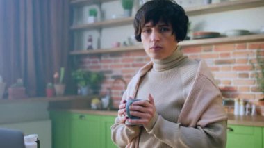 Mutfak adasındaki kameranın önünde kısa saçlı kadın işe başlamak için çok karizmatik bir şekilde çabalıyor ama kendini yarasa ve soğuk hissediyor elinde bir fincan çay tutuyor ve dizüstü bilgisayarına bakıyor. 4k