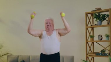 Evde, oturma odasında yaşlı ve karizmatik bir adam sağlıklı bir vücut için evde egzersiz yapmaktan çok mutlu. Pazılarını çalıştırıyor. 4k