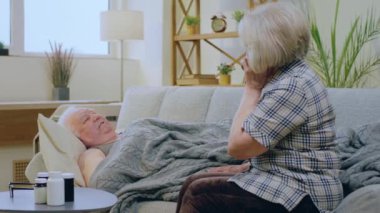 Hasta yaşlı adam oturma odasındaki kanepede yatıyor. Karısı tedavi için GP 'yi arıyor ya da sağlık kontrolü için randevu alıyor. 4k