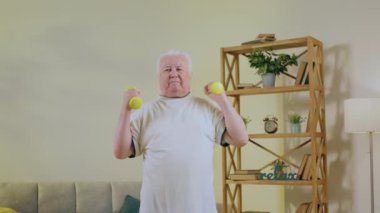 Kameraya yakın aydınlık bir oturma odasında mutlu ve heyecanlı yaşlı bir adam egzersiz yaparken kaslarını ağırlık kullanarak çalıştırıyor. 4k