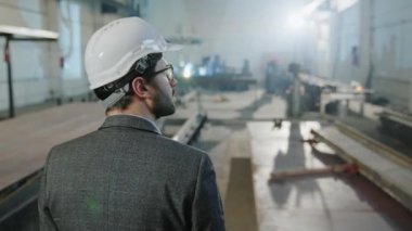 Üretim bölgesinin ortasında duran, çalışma planını tutan ve güvenlik kaskı taktığı durumu analiz eden bir yöneticinin arkasından video çekmek. 4k