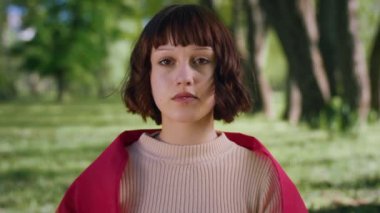 Kameraya yakın kısa saçlı bayan kameraya poz veriyor. Parkta kalıyor. Gözleri kapalı..