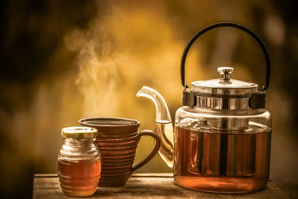 Çay demliği, çay fincanı ve ahşap bir masada bal olan cam bardak.