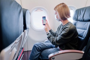 Yüz maskesi takan genç bir kadın akıllı telefon kullanarak uçakla seyahat ediyor. Yeni normal seyahat covid-19 salgın konsepti.