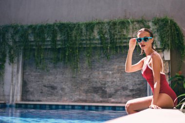 Bronzlaşmak ve tatilin tadını çıkarmak. Yüzme havuzunun yanında oturan kırmızı mayolu güzel bir kadın..