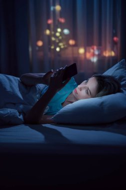 Genç, uykucu bir kadının portresi gece yarısı akıllı telefon kullanarak yatakta uzanıyor, uyuyamıyor. Uykusuzluk, uyku bozukluğu konsepti.