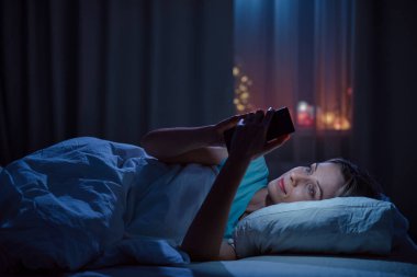 Geceleri rahat yatak odasında akıllı telefon kullanan genç bir kadın..