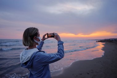 Medial maskeli bir kadın deniz sahilinde harika bir gün batımının fotoğrafını çekiyor..