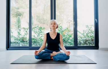 Evde, nilüferli yaşlı bir kadın büyük bir pencerenin önünde mindere oturmuş poz veriyor. Sakinlik ve meditasyon kavramı.