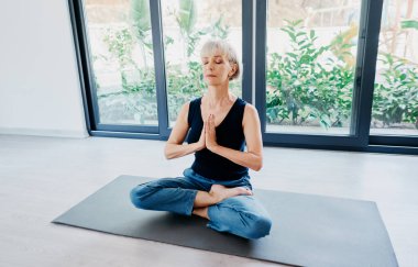 Evde, nilüferli yaşlı bir kadın büyük bir pencerenin önünde mindere oturmuş poz veriyor. Sakinlik ve meditasyon kavramı.