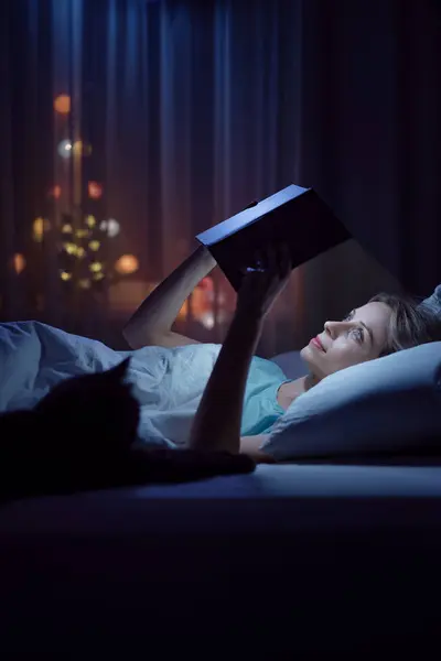 Magic night. Young woman reading book at cozy bed with her cat.