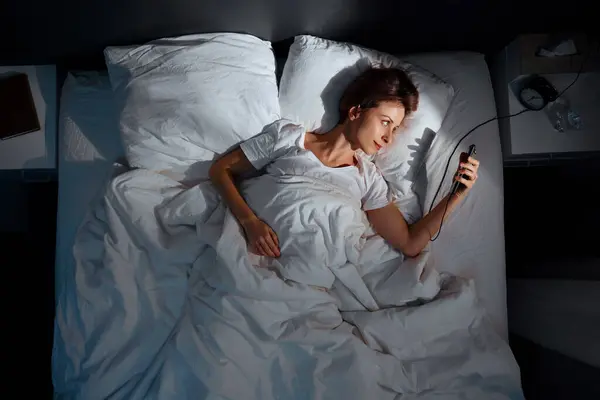 Young woman using smartphone in bed in her cozy bedroom at night. Top view.