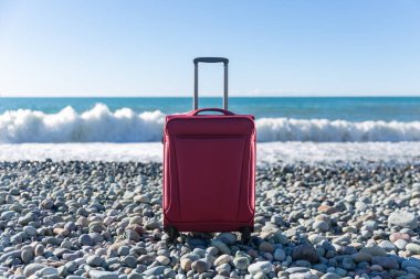 Color travel suitcase on pebble beach with turquoise sea background, summer holidays concept