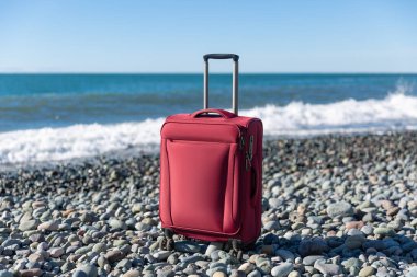 Color travel suitcase on pebble beach with turquoise sea background, summer holidays concept