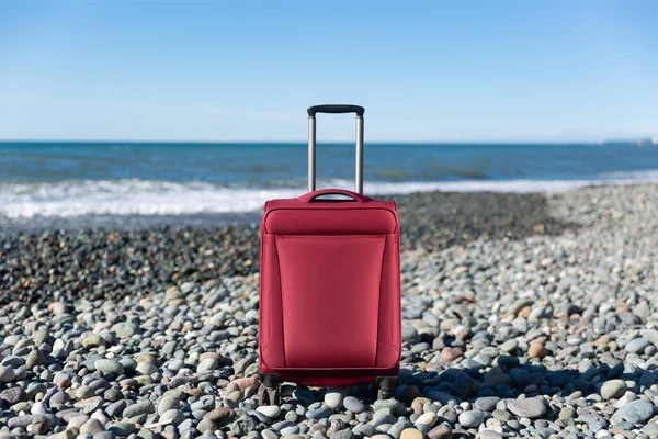 Color travel suitcase on pebble beach with turquoise sea background, summer holidays concept
