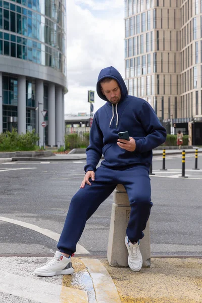 Full-length young man wears a blue sports suit and Looks at the phone. Athletic handsome young man sits on urban location and shows sports suit. Mock-up
