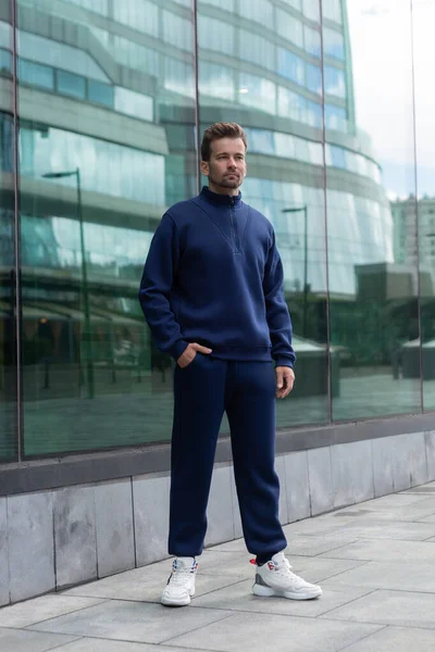 Full-length young man wears a vinous sports suit and poses for the camera. Athletic handsome young man stands on urban location and shows sports suit. Mock-up